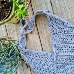 A handmade crochet envelope-style bag lying flat on weathered wooden decking. The bag is made in a light gray yarn with a textured chevron-like stitch pattern. It features a triangular fold-over flap design creating the envelope shape. The image includes partial views of a potted plant with long leaves and a yellow and red heart-patterned decorative item in the background. The bag's surface shows detailed stitch work in a uniform pattern, demonstrating careful craftsmanship in the crochet technique
