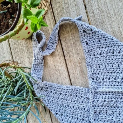 A handmade crochet envelope-style bag lying flat on weathered wooden decking. The bag is made in a light gray yarn with a textured chevron-like stitch pattern. It features a triangular fold-over flap design creating the envelope shape. The image includes partial views of a potted plant with long leaves and a yellow and red heart-patterned decorative item in the background. The bag's surface shows detailed stitch work in a uniform pattern, demonstrating careful craftsmanship in the crochet technique
