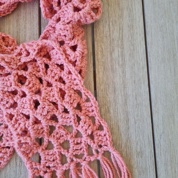 Coral pink lacy crochet scarf with shell pattern and long fringe displayed on white mannequin, featuring an airy diamond mesh design throughout