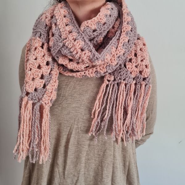 A handmade crochet scarf made from granny squares in soft pink and lavender tones, laid out on a wooden surface. The scarf features a delicate fringe along the edges, adding a cozy and stylish touch.