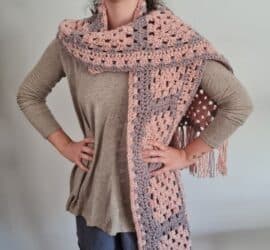 A handmade crochet scarf made from granny squares in soft pink and lavender tones, laid out on a wooden surface. The scarf features a delicate fringe along the edges, adding a cozy and stylish touch.