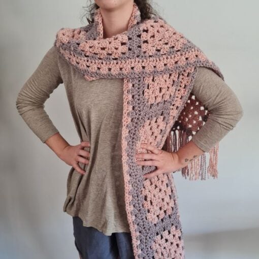 A handmade crochet scarf made from granny squares in soft pink and lavender tones, laid out on a wooden surface. The scarf features a delicate fringe along the edges, adding a cozy and stylish touch.