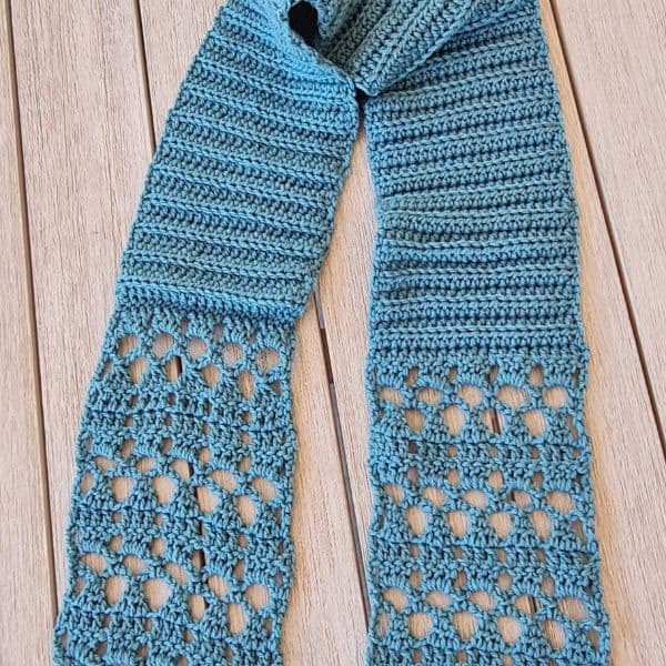 Light blue crochet scarf laid flat on wooden background, featuring ribbed texture at neck transitioning into delicate diamond lace pattern at both ends. Pull-through style scarf showcasing intricate stitch work