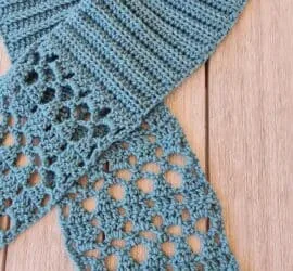 Light blue crochet scarf laid flat on wooden background, featuring ribbed texture at neck transitioning into delicate diamond lace pattern at both ends. Pull-through style scarf showcasing intricate stitch work