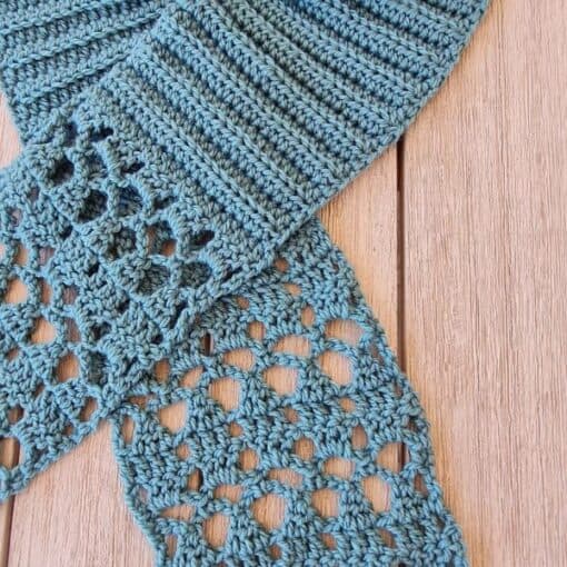 Light blue crochet scarf laid flat on wooden background, featuring ribbed texture at neck transitioning into delicate diamond lace pattern at both ends. Pull-through style scarf showcasing intricate stitch work