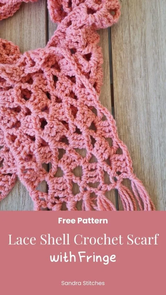 Coral pink lacy crochet scarf with shell pattern and long fringe displayed on white mannequin, featuring an airy diamond mesh design throughout