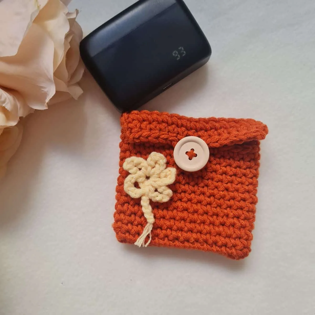 lat lay of a small burnt orange crochet pouch featuring a unique yarn under 'X' stitch texture, a yellow floral appliqué, and a light wood button closure.