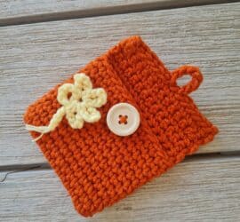 lat lay of a small burnt orange crochet pouch featuring a unique yarn under 'X' stitch texture, a yellow floral appliqué, and a light wood button closure