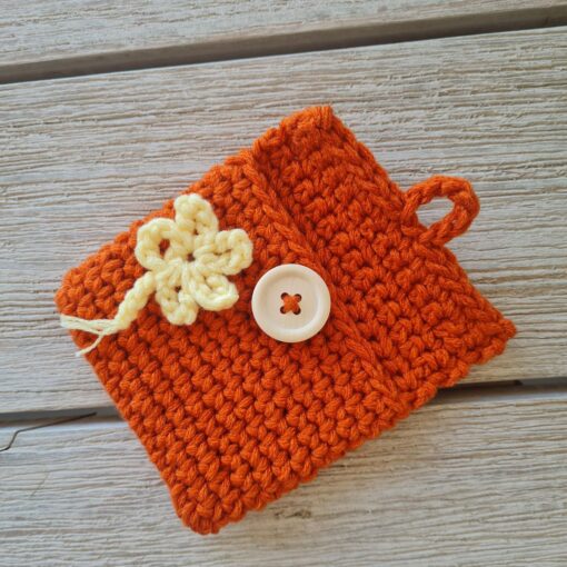 lat lay of a small burnt orange crochet pouch featuring a unique yarn under 'X' stitch texture, a yellow floral appliqué, and a light wood button closure