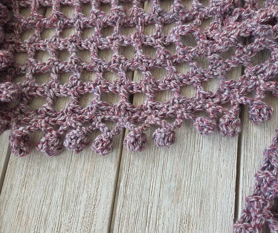 A row of small crocheted pompoms along the back hem of a crochet mesh shrug.