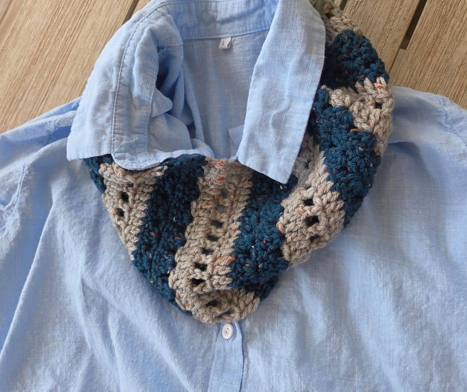 A striped crochet cowl in deep teal and flecked grey, worked in textured bands and displayed on a light blue shirt