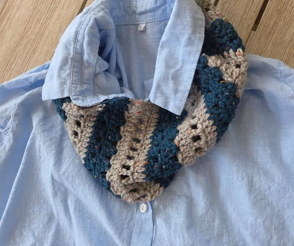A striped crochet cowl in deep teal and flecked grey, worked in textured bands and displayed on a light blue shirt